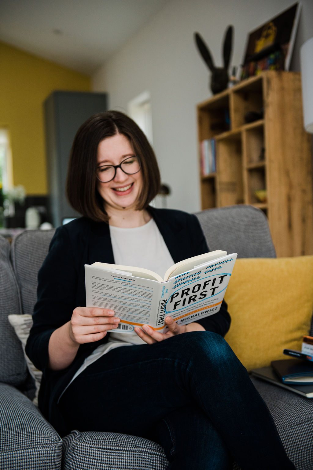 Jenny Pace, a 30-something professional woman, is sat on a grey sofa reading a book named Profit First. She is leaning on a yellow cushion and smiling.